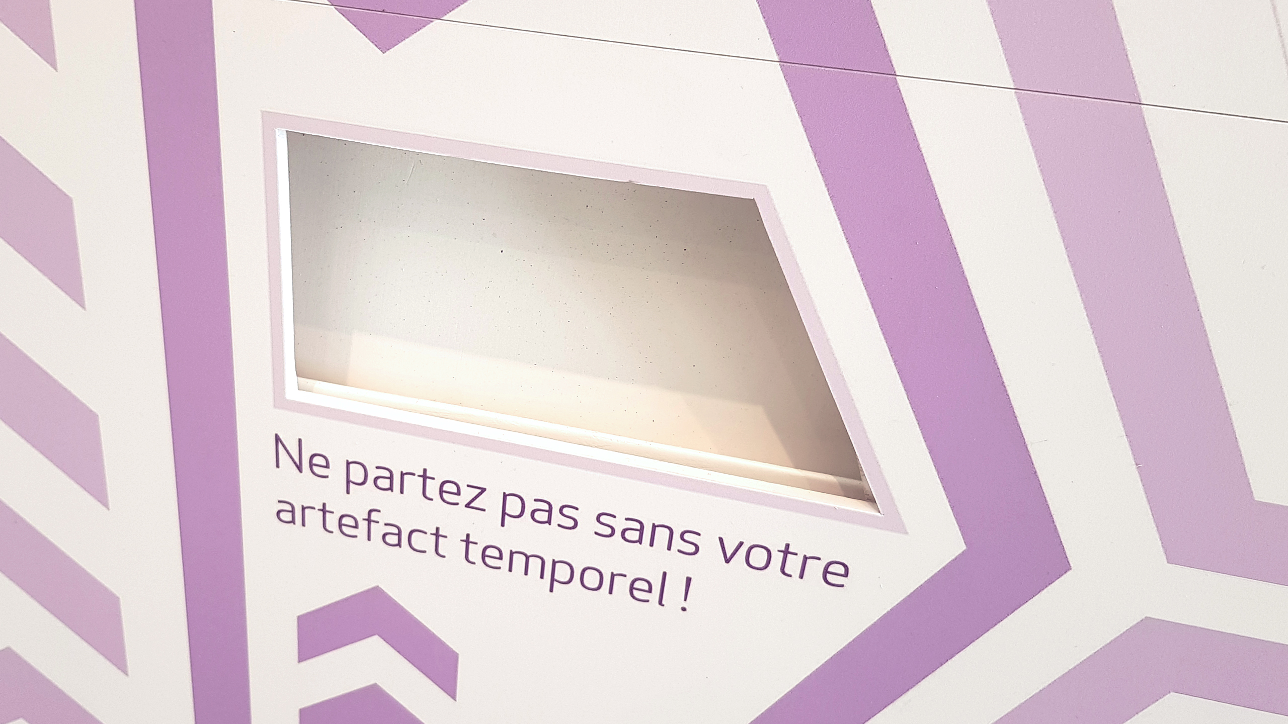 Photographie en gros plan de l'ouverture à l'extérieur de la cabine permettant de récupérer son illustration. Un message est présent : "Ne partez pas sans votre artefact temporel !"