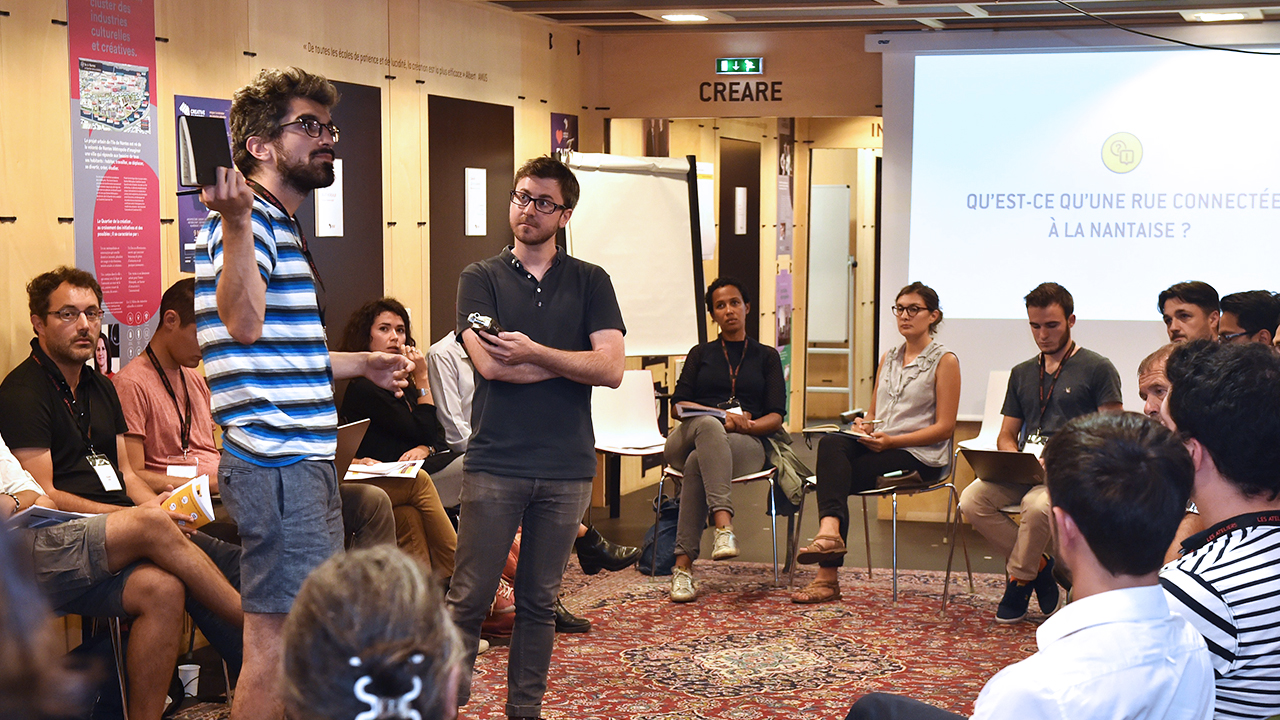 Photographie d'un atelier. Les participants sont assis en cercle, autour de l'un d'entre eux qui prend la parole, debout au centre du cercle. Un animateur est présent à côté de lui. En arrière-plan figure une diapositive titrée "Qu'est-ce qu'une rue connectée à la nantaise ?"