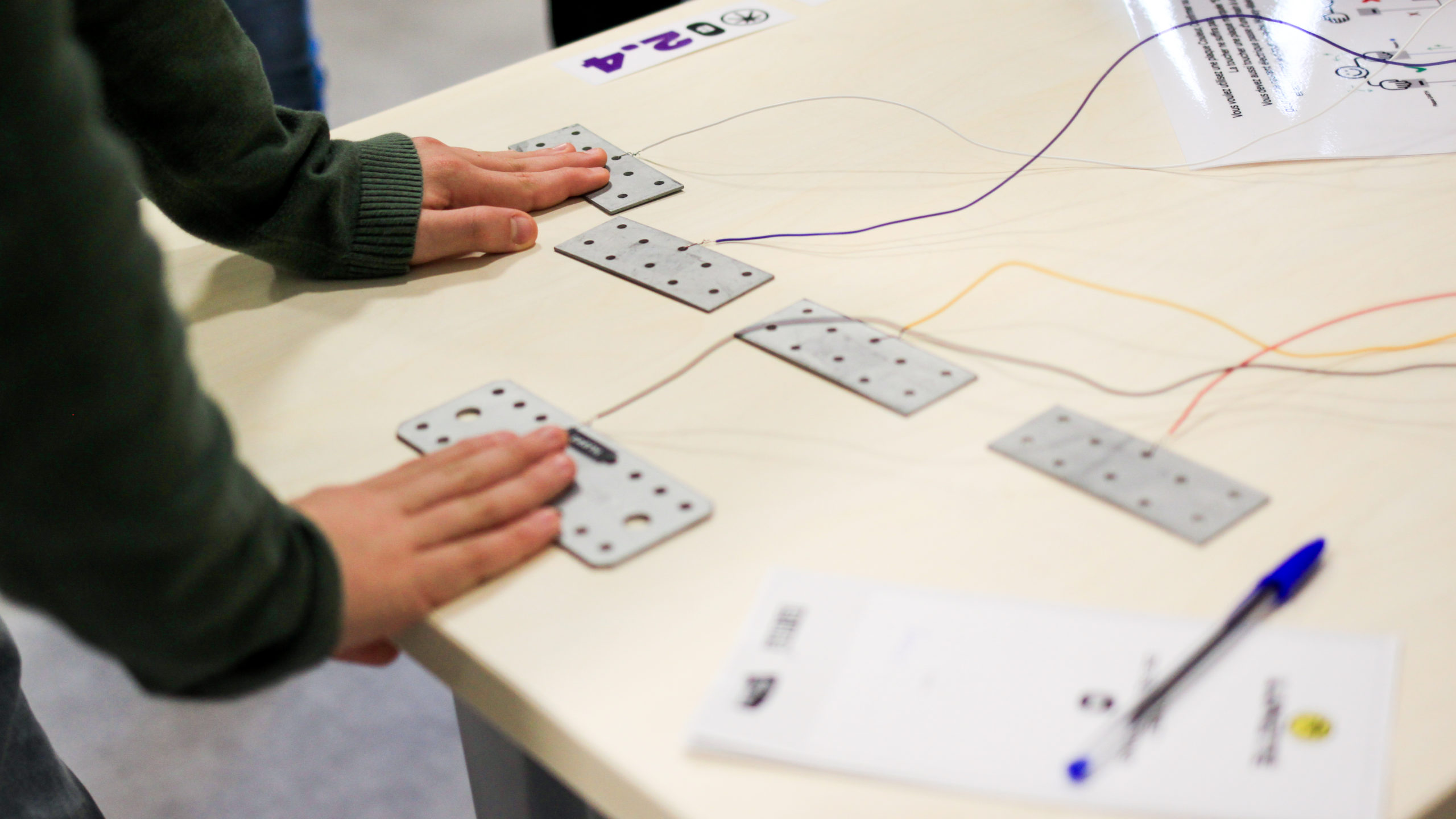 Photographie en gros plan sur des plaques métalliques placées sur une table, reliées par des fils conducteurs à un dispositive en dehors du cadre. Un participant touche plusieurs plaques dans le but de résoudre une énigme.