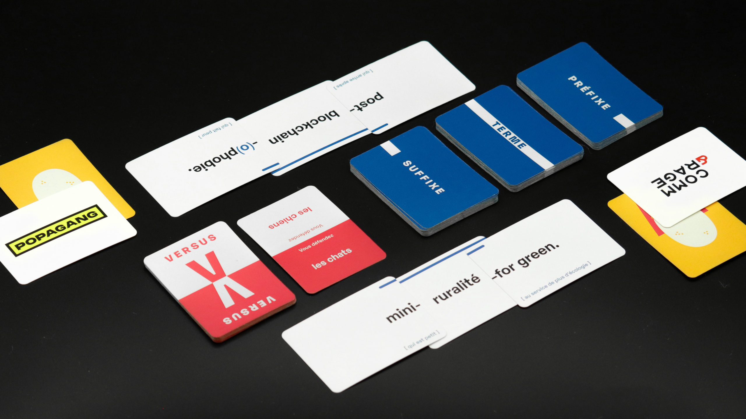 Photographie d'une mise en place du jeu. 3 paquets "Préfixe", Terme" et "Suffixe", ainsi qu'un paquet "Versus" sont placés au centre. Une carte Versus est retournée et indique d'une part "Vous défendez les chats", et d'autre "Vous défendez les chiens". Un des joueurs a tiré de son côté 3 cartes formant le néologisme "post-blockchain-ophobie". L'autre joueur a tiré des cartes formant le néologisme "mini-ruralité-for green".